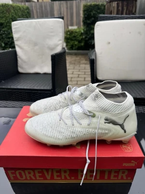 Vita Puma fotbollsskor med strumpkrage - Snygga vita elite fotbollsskor från Puma med stickad strumpkrage för extra stöd runt ankeln. Skorna har en modern design med grå detaljer och Puma-logga på sidan. Ovandelen är tillverkad i syntetmaterial och snörningen ger bra passform. Perfekta för dig som vill sticka ut på planen. Använt endast en gång! Nypris 2700 kr.