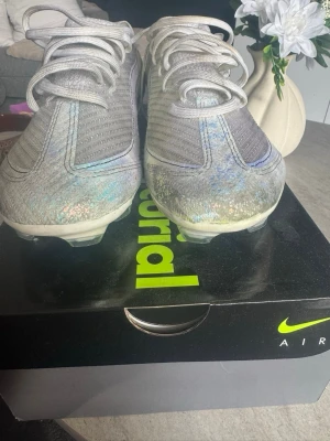 Nike Mercurial silvriga fotbollsskor - Säljer ett par Nike Mercurial fotbollsskor i silver med skimrande detaljer. Skorna har snörning, låg profil och är tillverkade i ett lätt syntetmaterial som ger bra bollkontroll. Perfekta för dig som vill sticka ut på planen!