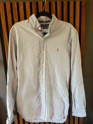 Ljusblå skjorta från Polo Ralph Lauren - Klassisk ljusblå skjorta från Polo Ralph Lauren med broderad logga på bröstet. Skjortan har knappar framtill och button-down krage. Perfekt för en stilren look och tillverkad i mjuk bomull.