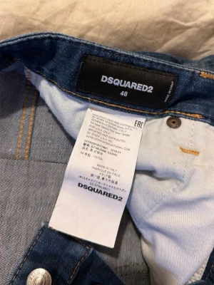 Blå jeans från Dsquared2, storlek 48 - Säljer ett par snygga blå jeans från Dsquared2 i storlek 48. Jeansen har klassisk femficksmodell, slitna detaljer och kontrastsömmar. Knapparna framtill har Dsquared2-logga och det finns en röd logotagg vid gylfen. Tillverkade i Italien.