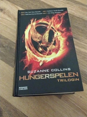 Hungerspelen Trilogin av Suzanne Collins - Hungerspelen Trilogin av Suzanne Collins i en samlingsvolym. Omslaget är svart med ett brinnande gyllene emblem och text i guld och vitt. Innehåller Hungerspelen, Fatta eld och Revolt. Perfekt för dig som gillar dystopiska äventyr och spänning.