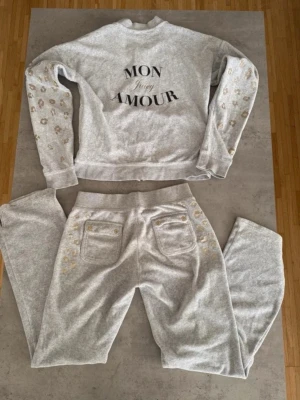 Vintage Juicy Couture sweatshirtset - Mysigt grått sweatshirtset från Juicy Couture med texten 'MON AMOUR' på tröjan och leopardmönster i beige och guld på ärmar och byxor. Setet består av en sweatshirt och en ziptröja, båda med långa ärmar och ribbade muddar. Perfekt för chill dagar. 