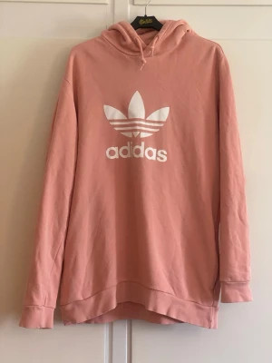 Rosa hoodie från Adidas med logga - Säljer en snygg rosa hoodie från Adidas med stor vit Trefoil-logga och text på bröstet. Hoodien har huva med dragsko och långa ärmar. Tillverkad i mjukt bomullsmaterial, perfekt för en avslappnad streetwear-look.