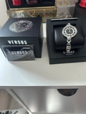 Versus Versace silverfärgad klocka - Stilren klocka från Versus Versace i silverfärg med svart urtavla. Klockan har ett runt boett med dekorativa detaljer och länkar, samt tydliga bokstäver runt urtavlan. Perfekt accessoar för dig som gillar exklusiv design.