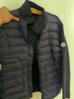 Mörkblå  Moncler Cardigan - Snygg mörkblå pufferjacka från Moncler med quiltad front och stickade ärmar. Jackan har hög krage, dragkedja framtill och Moncler-logga på ena ärmen. Perfekt för kalla vinterdagar och ger en clean, modern look.