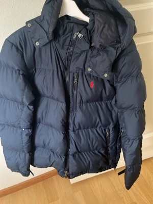Mörkblå pufferjacka från Polo Ralph Lauren i storlek S - Snygg mörkblå pufferjacka från Polo Ralph Lauren med huva och dragkedja. Jackan har en röd logga på bröstet, flera fickor med dragkedja och en quiltad design som håller värmen. Perfekt för kalla vinterdagar och riktigt stilren. Nypris minst 3000 kr