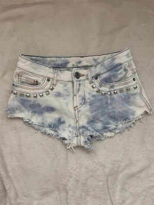 Tie-dye shorts med nitar - Säljer ett par vita och ljusblå tie-dye shorts med råa kanter och silverfärgade nitar vid fickorna. Shortsen har fem fickor och knappgylf. Perfekta för dig som vill ha en edgy och trendig stil. Storlek saknas men uppskattas xs/s😇