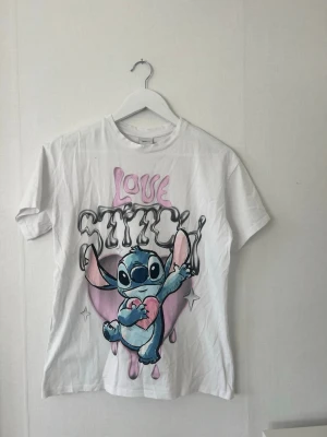 Vit Disney Stitch T-shirt - Vit T-shirt från Disney med stort tryck av Stitch och texten 'Love Stitch' i rosa och svart. T-shirten har korta ärmar, rund halsringning och är gjord i mjuk bomull. Perfekt för dig som älskar söta och färgglada motiv! Står ej storlek men upplever den som en S