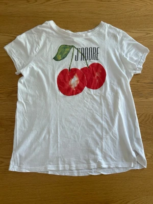 Vit t-shirt med körsbär och text - Vit t-shirt i bomull med tryck av två stora röda körsbär och texten 'J'ADORE' på bröstet. Klassisk rund hals och korta ärmar. Perfekt för dig som gillar färgglada prints och vill ha en chill look.