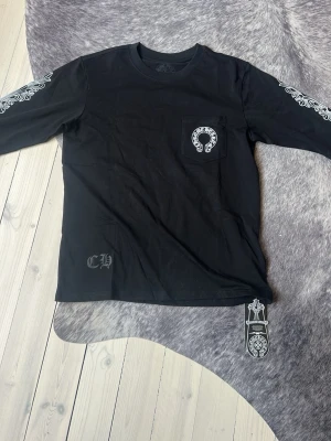 Svart långärmad topp från Chrome Hearts - Svart långärmad topp från Chrome Hearts med tryck på bröstfickan och ärmarna. Snygga vita detaljer och logga på bröstet samt coolt mönster längs båda ärmarna. Tillverkad i mjuk bomull för en bekväm känsla.