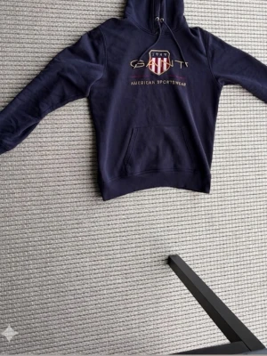 Marinblå hoodie från Gant - Snygg marinblå hoodie från Gant med broderad logga och texten 'American Sportswear' på bröstet. Klassisk känguruficka framtill och justerbar huva med snörning. Perfekt för en avslappnad och sportig stil. Storlek L i bra skick.