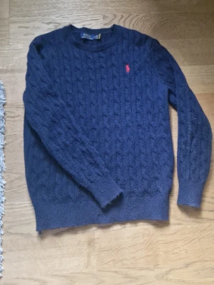 Mörkblå kabelstickad tröja Polo Ralph Lauren - Mörkblå kabelstickad tröja från Polo Ralph Lauren med rund halsringning och röd broderad logga på bröstet. Tröjan har ribbade muddar vid ärmslut och nederkant. Klassisk och tidlös design i mjukt material.
