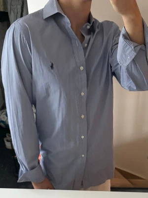 Blårandig skjorta från Ralph Lauren - Klassisk blårandig skjorta från Ralph Lauren med vit knappstängning och mörkblå broderad logga på bröstet. Skjortan har lång ärm, krage och är tillverkad i bomull. Perfekt för en clean och stilren look. Storlek 39 vilket motsvarar m ungefär 