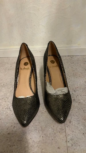 Svarta pumps med ormmönster La Strada - Snygga svarta pumps från La Strada med trendigt ormmönster och spetsig tå. Skorna har en elegant form och en klassisk klack. Utsidan har en glansig finish som ger extra attityd till outfiten. Perfekta för dig som vill sticka ut lite extra.