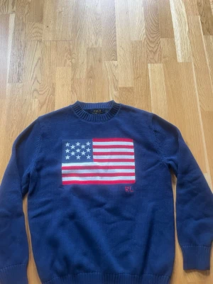 Blå stickad tröja Polo Ralph Lauren - Mörkblå stickad tröja från Polo Ralph Lauren med amerikansk flagga i rött, vitt och blått på bröstet. Rund halsringning och ribbade muddar. RL-broderi i rött nedtill på flaggan. Perfekt för dig som gillar klassisk preppy stil.