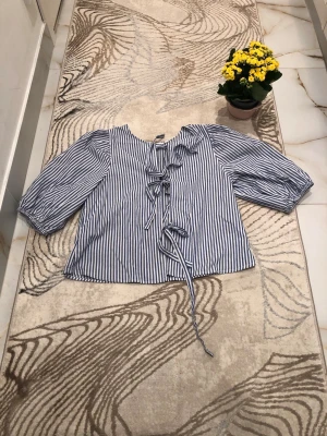 Randig blus med knytning från Kappahl - Snygg blus med blåvita vertikala ränder och trekvartsärm. Framtill har den flera dekorativa knytband och en liten volang vid halsen. Tillverkad i ett lätt bomullsliknande material som känns luftigt och fräscht. Använd endast en gång. Är öppen för prisförslag😊