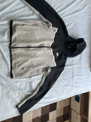 Nike grå och svart hoodie med dragkedja - Nike hoodie i grått och svart med hel dragkedja framtill och vit logga på bröstet. Hoodien har huva, två sidofickor och en extra ficka med dragkedja på ena ärmen. Tillverkad i mjukt material, perfekt för en sportig och avslappnad stil. 