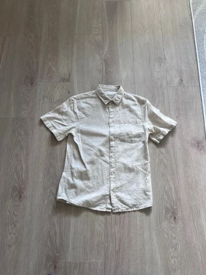 Beige linneskjorta från HM - Snygg beige skjorta med korta armar och klassisk krage. Skjortan har en bröstficka och knäppning framtill. Perfekt för en avslappnad och stilren look.