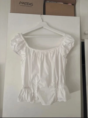 Vit blus med puffärm och volang - Söt vit blus med puffärmar och volang nedtill. Blusen har en bred, rundad urringning och resår vid ärmslut och midja för en feminin siluett. Perfekt till jeans eller kjol för en trendig look.