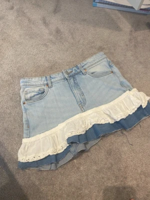 Ljus jeanskjol med volangdetalj - Supersöt kort jeanskjol i ljusblå denim med volang i vitt tyg längst ner. Kjolen har klassiska fickor fram och bak, bälteshällor och knäppning framtill. Volangen ger kjolen en extra feminin touch och gör den unik.