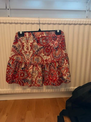 Röd paisleymönstrad volangkjol - Kort kjol med volang och paisleymönster i rött, blått och beige. Kjolen har en avslappnad passform och resår i midjan för extra komfort. Perfekt för dig som gillar färgstarka och bohemiska plagg.