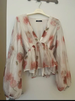 Blommig blus med ballongärmar Gina Tricot - Romantisk blus från Gina Tricot i transparent vitt tyg med rosa och gröna blommor. Djup v-ringning, markerad midja och volangkant nedtill. Långa ballongärmar ger en luftig och trendig look. Perfekt att styla med jeans eller kjol.