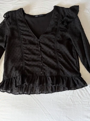 Svart blus med volang från Zara - Snygg svart blus från Zara med diskret prickigt mönster, volangdetaljer vid axlar och nederkant samt v-ringning och knappar framtill. Tillverkad i ett tunt, lätt material som ger en luftig känsla. Perfekt för att lyfta vilken outfit som helst. Köpare står för frakt. 