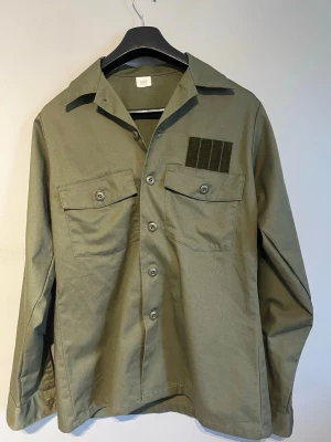 Olivgrön, vintage militärjacka/overshirt med fickor - Vintage, olivgrön militärjacka/ overshirt med två stora bröstfickor och knappstängning framtill. Eftersom den är en äkta vintage militärjacka är den unik och ganska rare. Overshirten har klassisk krage och dekorativ detalj på ena fickan. Tillverkad i ett slitstarkt bomullstyg. Jackan är tunn och sval. Perfekt för sommar, vår och höst för lager på lager och en avslappnad look. Tror den är insydd lite i midjan men är inte 100 då den är vintage, men det påverkar inte passformen mycket alls. 