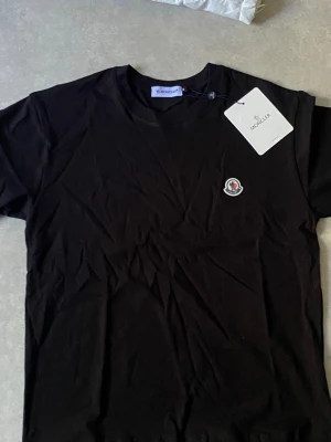 Svart Moncler t-shirt med logga - Snygg svart t-shirt från Moncler med klassisk rund hals och liten Moncler-logga på bröstet. T-shirten är kortärmad och har en enkel, stilren design som passar till det mesta. Tillverkad i mjuk bomull för skön känsla.