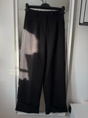Vida svarta kostymbyxor - Säljer ett par svarta kostymbyxor med vida ben och uppvikta benslut. Byxorna har hög midja, bälteshällor och pressveck framtill för en clean look. Perfekta till en trendig outfit och passar till många olika stilar.