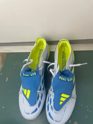 Adidas Predator fotbollsskor blå/gul elite - Säljer ett par Adidas Predator fotbollsskor i vitt, blått och neongult. Skorna har klassisk snörning, täckt plös med texten 'EST. 1994' och metallskruvdobbar för bästa grepp på planen. Snygg design med moderna detaljer och Predator-logga på sulan.