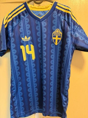 Sverige fotbollströja Adidas blå - Blå Sverige fotbollströja från Adidas med gula detaljer, nummer 14 och namnet Gyökeres på ryggen. Tröjan har klassiskt mönster, korta ärmar och gulblå ränder på axlarna. Perfekt för dig som vill stötta landslaget på match eller träning.