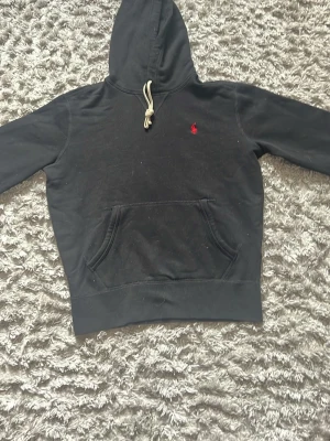 Svart hoodie från Polo Ralph Lauren - Klassisk svart hoodie från Polo Ralph Lauren med röd broderad logga på bröstet. Hoodien har känguruficka, huva med beige dragsko och ribbade muddar. Perfekt för en avslappnad och stilren look.