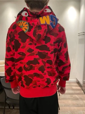 Röd camo hoodie från BAPE - Säljer en röd camo hoodie från BAPE med ikoniskt hajmotiv på huvan och broderade bokstäver. Hoodien har dragkedja framtill, ribbade muddar och detaljer i svart och brunt. Perfekt för dig som vill sticka ut med streetwear-stil.