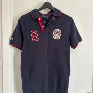 Mörkblå pikétröja från Tommy Hilfiger - Snygg mörkblå pikétröja från Tommy Hilfiger med röd och vit detaljering. Kortärmad med klassisk krage och knappar framtill. Perfekt för en avslappnad stil. Nypris 1100kr