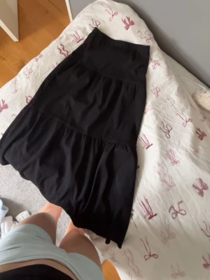 Svart långkjol med volanger - Säljer en svart långkjol med volangdetaljer och lätt rynkad struktur. Kjolen har en enkel och flowy design som passar perfekt till många olika stilar. Materialet känns mjukt och bekvämt mot huden.