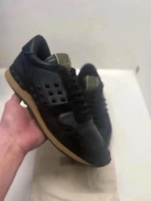 Svarta Valentino rockrunners  - Snygga svarta sneakers från Valentino med coola pyramidformade nitar på sidorna och olivgröna detaljer på plösen. Skorna har en klassisk låg siluett och gummisula i kontrasterande beige. Materialmix av läder och textil ger en lyxig känsla.