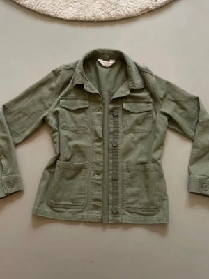 Grön utilityjacka från H&M - Snygg grön utilityjacka från H&M med fyra stora fickor framtill och knappar längs hela framsidan. Jackan har klassisk krage och är tillverkad i bomull, perfekt för lager på lager under vår och höst.