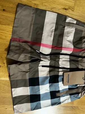 Rutig Burberry shorts med snörning - Säljer ett par klassiska Burberry shorts med rutigt mönster i beige, svart, vit och röd. De har snörning i midjan och är tillverkade i ett lätt syntetmaterial. Perfekta för sommarens alla äventyr och har den ikoniska Burberry-looken. Storlek M.