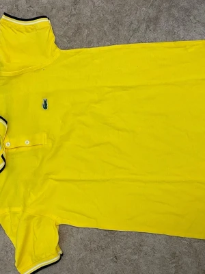 Gul pikétröja från Lacoste - Snygg gul pikétröja från Lacoste med klassisk krage och två knappar framtill. Tröjan har korta ärmar och den ikoniska gröna krokodilloggan på bröstet. Svarta och vita detaljer på krage och ärmslut ger en sportig touch.