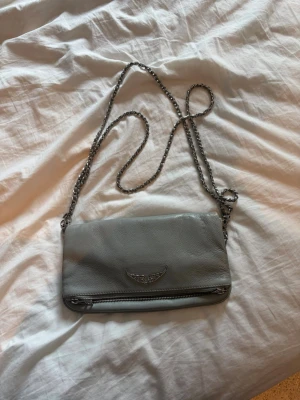 Grå axelväska från Zadig & Voltaire - Snygg grå axelväska i mjukt läder från Zadig & Voltaire. Väskan har en silverfärgad kedjerem och ett coolt emblem med vingar framtill. Dragkedja längst ner på framsidan för extra detaljer. Perfekt för dig som gillar stilrena accessoarer.