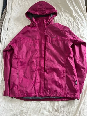 Rosa vindjacka med huva från Didriksons - Snygg rosa vindjacka från Didriksons med justerbar huva och dragkedja framtill. Jackan har två sidofickor med dragkedja och diskret logga på bröstet. Tillverkad i lätt och slitstarkt material, perfekt för blåsiga dagar. Jackan har storleken 160 men funkar även för vuxna kvinnor som är lite mindre.