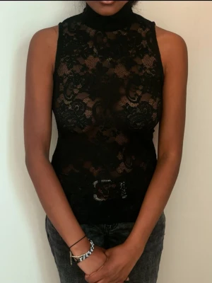 Svart spetsig polotopp - Säljer en svart ärmlös polotopp i genombruten spets med blommigt mönster. Toppen är tight och har hög hals, perfekt för att skapa en edgy look. Materialet är mjukt och stretchigt syntet. Snygg att styla med jeans eller kjol.
