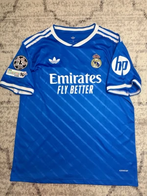 Blå Real Madrid matchtröja Adidas - Snygg blå Real Madrid matchtröja från Adidas med vita detaljer och klubbmärke på bröstet. Tröjan har vita ränder på axlarna, sponsorloggor och patchar på ärmarna. Tillverkad i lätt och ventilerande material, perfekt för fotboll eller träning.