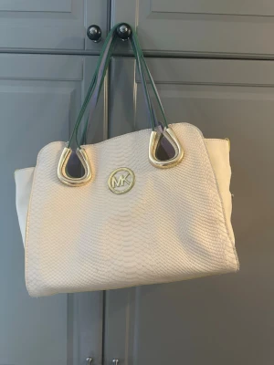 Vit MK inspirerade  handväska i ormmönster - Snygg vit handväska från Michael Kors med ormmönstrad struktur och guldfärgade detaljer. Väskan har gröna och lila axelband samt ett stort MK-emblem framtill. Innehåller en löstagbar innerväska i leopard mönster. Perfekt för dig som vill ha en trendig och rymlig väska med lyxig känsla.