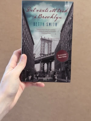 Det växte ett träd i Brooklyn - Romanen 'Det växte ett träd i Brooklyn' av Betty Smith är en klassiker med ett svartvitt omslag som visar en ikonisk bro och stadsmiljö. Boken är i pocketformat och har tydliga titeldetaljer i rött och blått. Perfekt för dig som gillar gripande berättelser om livet.