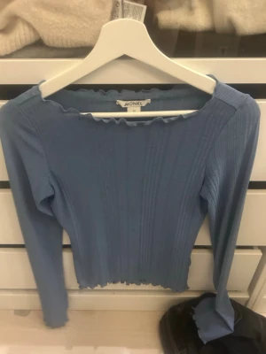 Blå ribbad topp från Monki - Supersöt blå långärmad topp från Monki med ribbad struktur och vågig kant vid halsringning och nederkant. Perfekt att styla med jeans eller kjol för en trendig look. Toppens material känns mjukt och stretchigt.