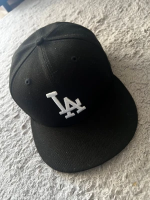 Svart LA-keps från New Era - Säljer en svart keps från New Era med LA-logga broderad i vitt framtill. Klassisk rak skärm och ventilerande hål upptill. Perfekt för dig som gillar streetwear och sportig stil. Materialet är tåligt och kepsen är helt nyköpt