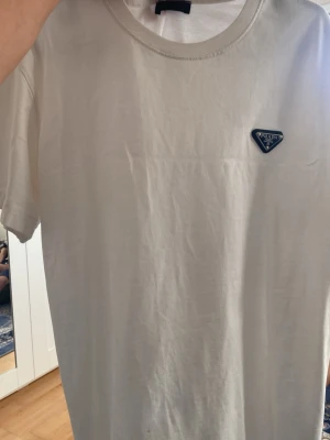 Vit t-shirt från Prada med logga - Säljer en stilren vit t-shirt från Prada med klassisk rund hals och korta ärmar. På bröstet sitter Pradas ikoniska trekantiga logga i svart och silver. Tillverkad i mjuk bomull för en skön känsla och clean look.