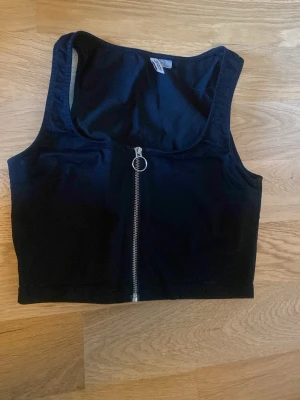 Svart croppad topp med dragkedja - Snygg svart croppad topp med rund halsringning och en silverfärgad dragkedja framtill. Perfekt att styla till jeans eller kjol för en trendig look. Toppen är ärmlös och har en sportig vibe.